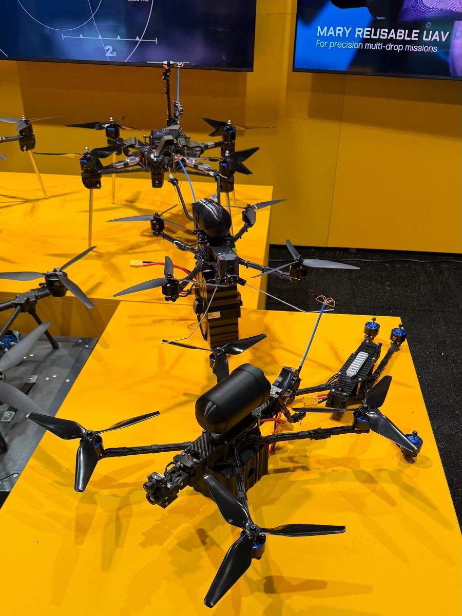 FPV and multi-rotor drones displayed on a yellow stand, with a screen reading reusable UAV for precision multi-drop missions