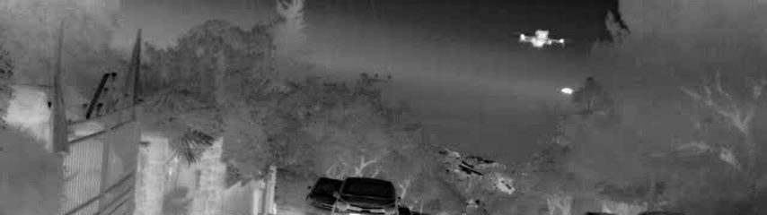 Thermal image of a quadcopter drone in flight above trees and parked vehicles